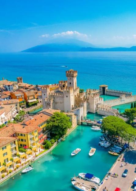 Castello medievale sul lago con barche, circondato da edifici colorati e natura rigogliosa.