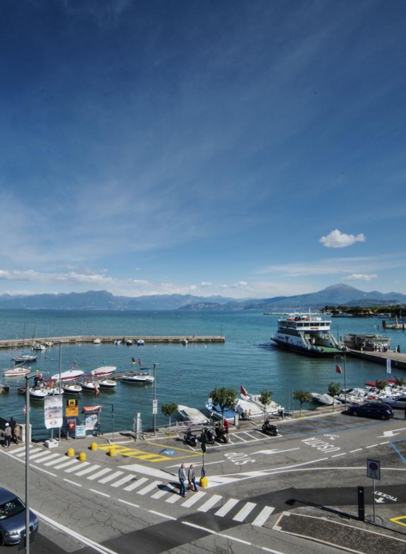 Vista del porto con barche e traghetto, cielo sereno e montagne sullo sfondo.