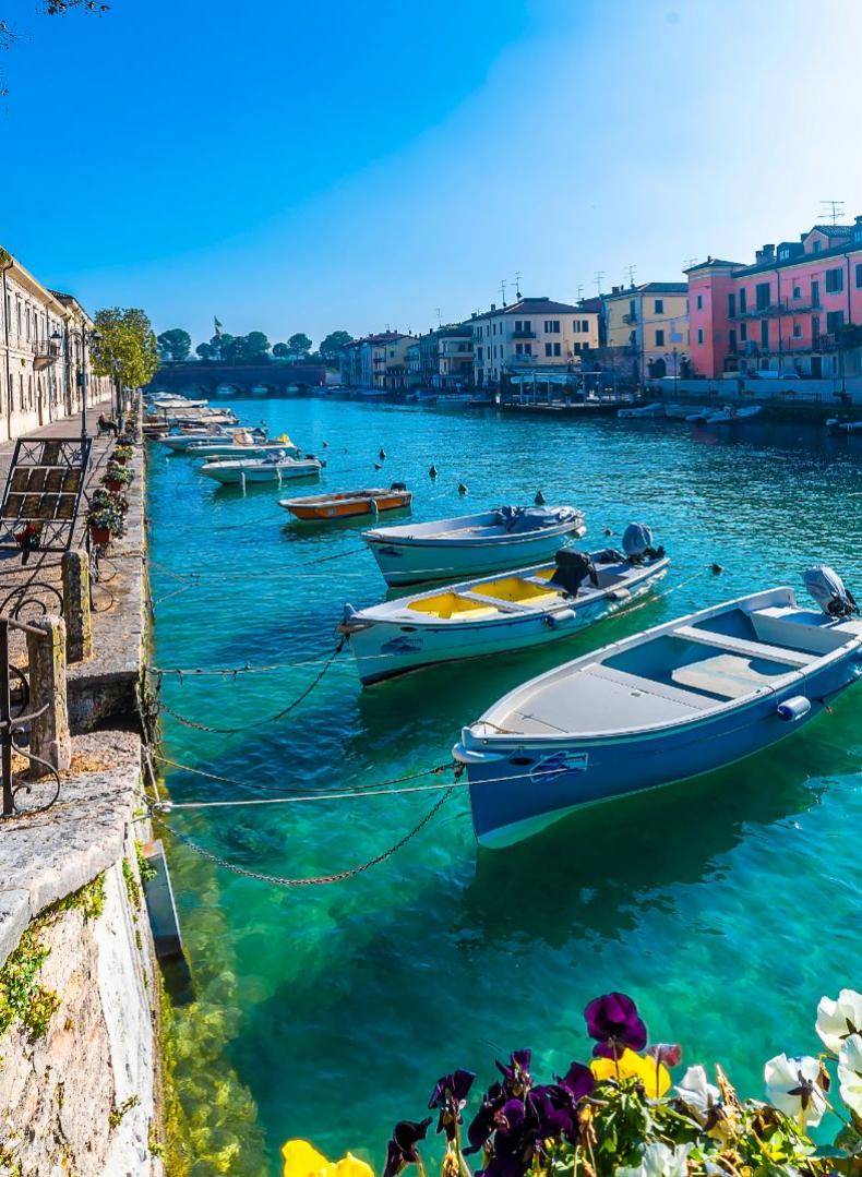 Barche ormeggiate lungo un canale con edifici colorati e fiori in primo piano.