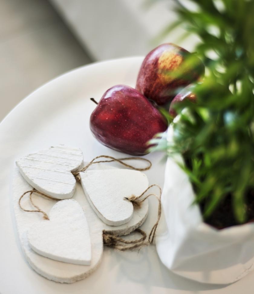 Tavolo bianco con mele rosse, cuori di legno e pianta verde.