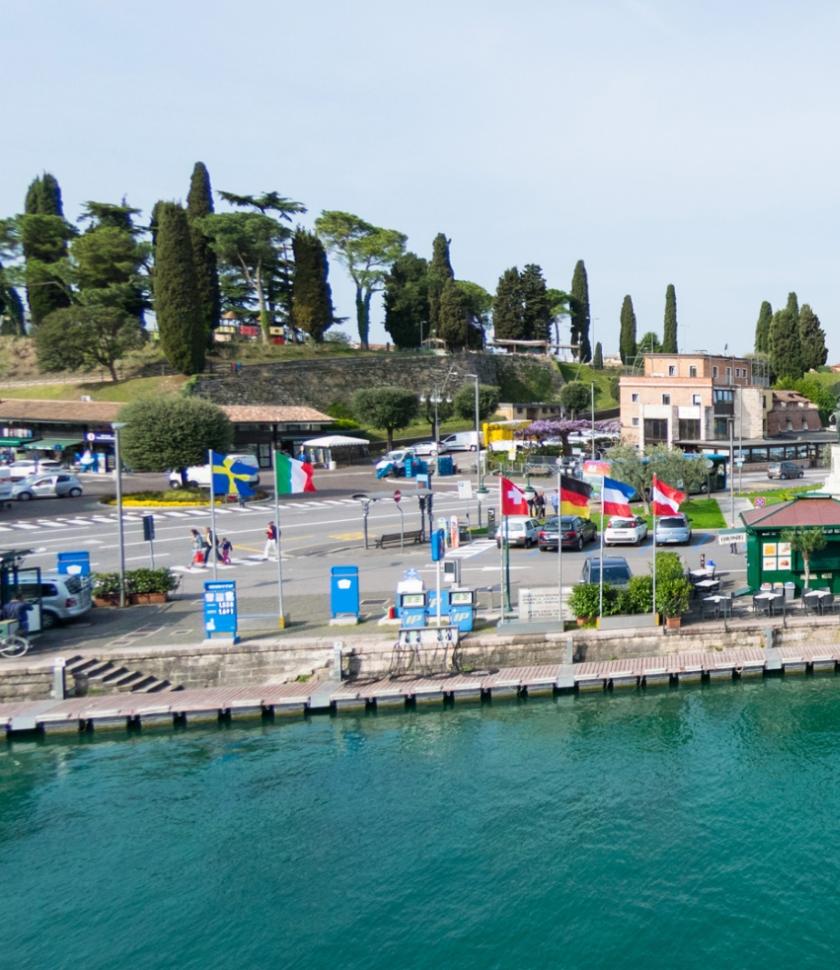 Lungolago con bandiere, parcheggio e verde, vicino a un porto turistico.