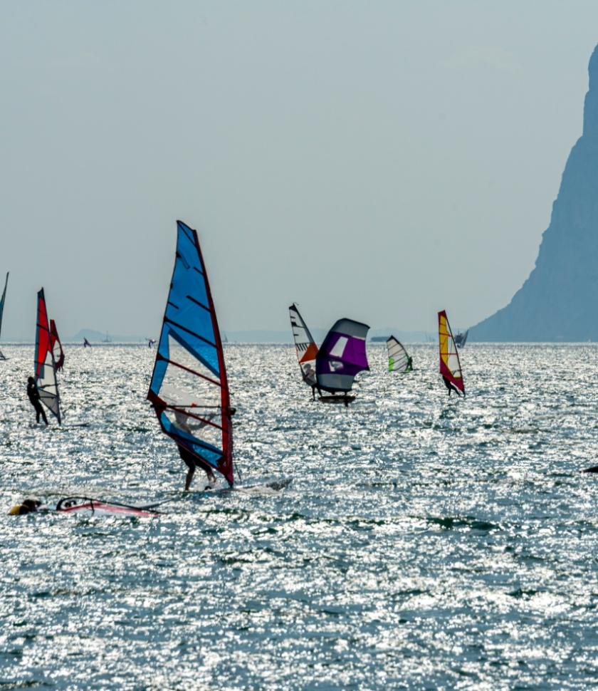 Windsurfisti colorati solcano il mare scintillante, con montagne sullo sfondo.