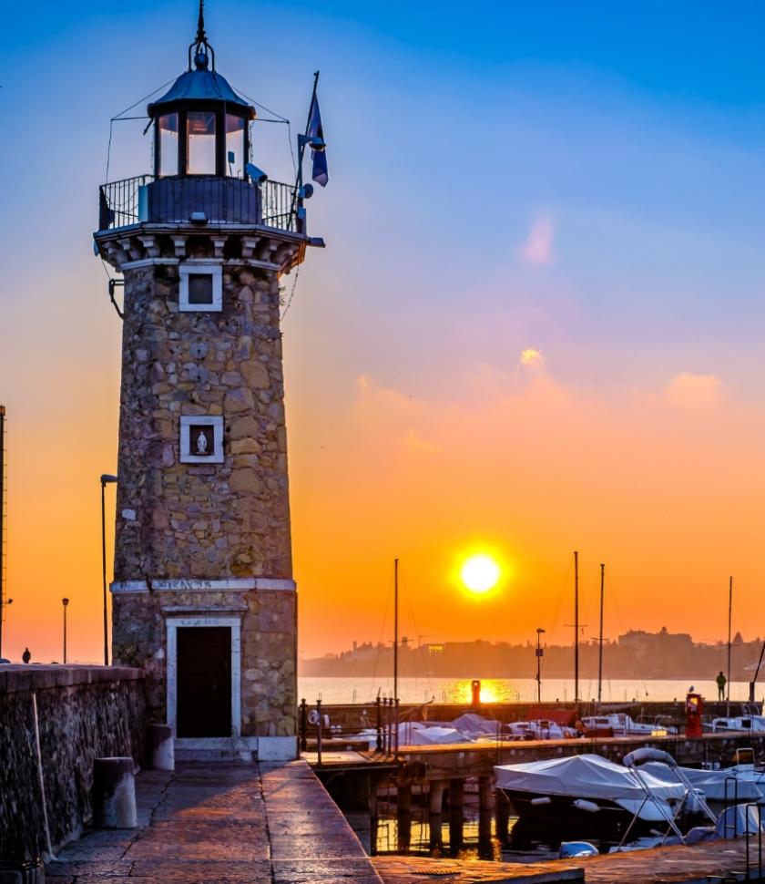 Faro al tramonto con barche ormeggiate, cielo arancione e blu.